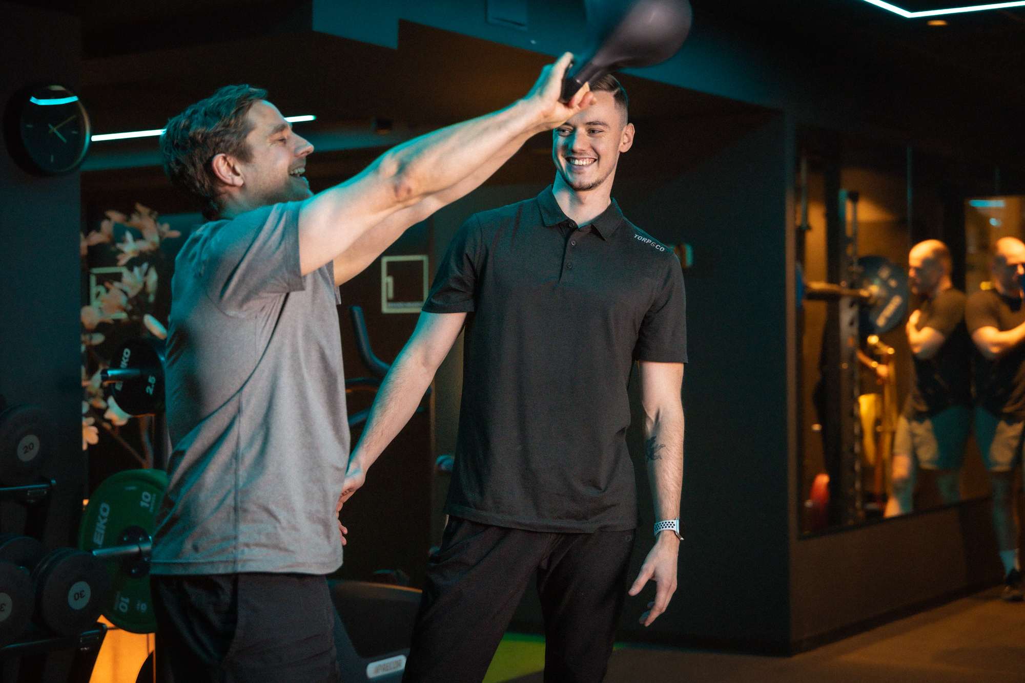 Mann løfter kettlebell over hodet hos Torp&Co mens en trener følger med.