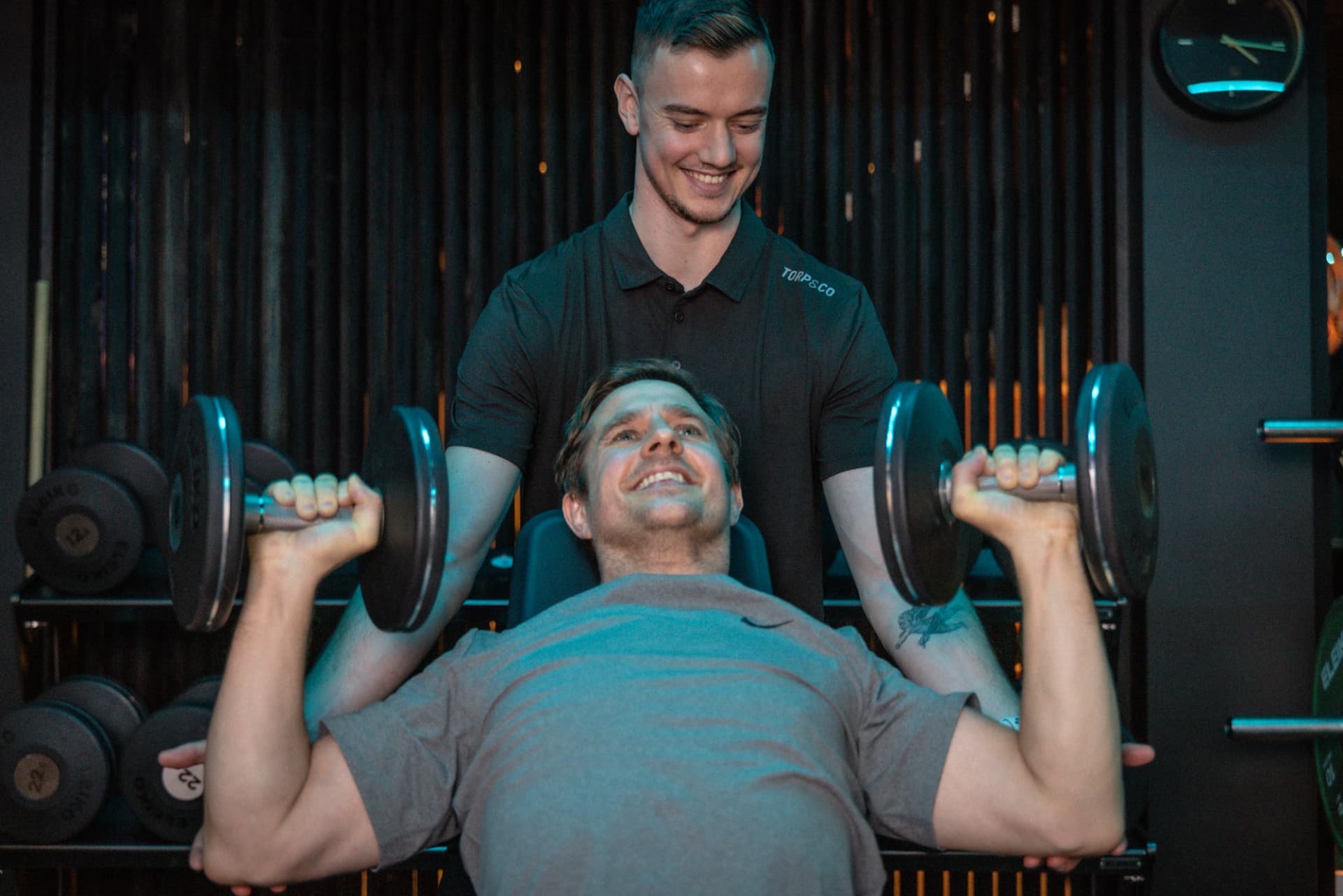 Mann utfører incline dumbbell press på skrå benk hos Torp&Co med trener som spotter.